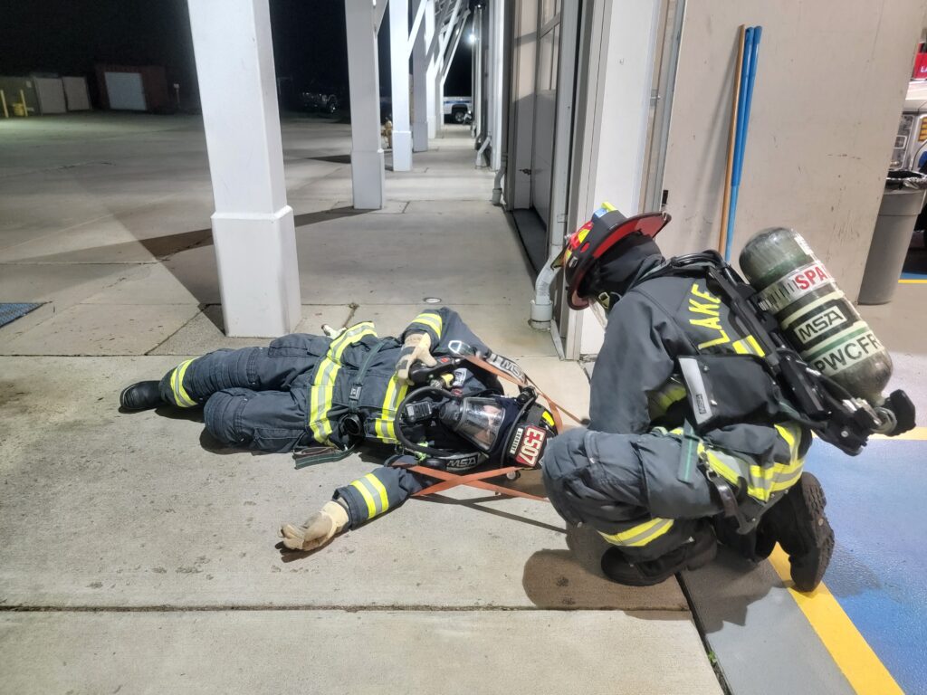 A firefighter in full gear assists or checks on another firefighter lying on the ground outside a building at night.