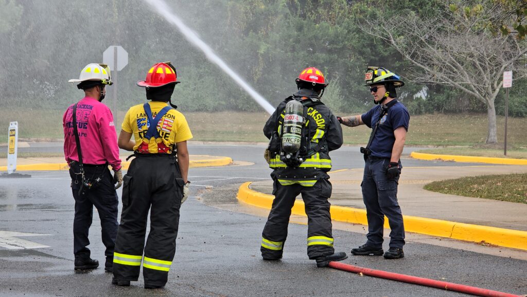Four firefighters in gear stand on a wet street, with one spraying water from a hose. Trees and yellow curbs are in the background. The scene appears to be a training exercise or emergency response.