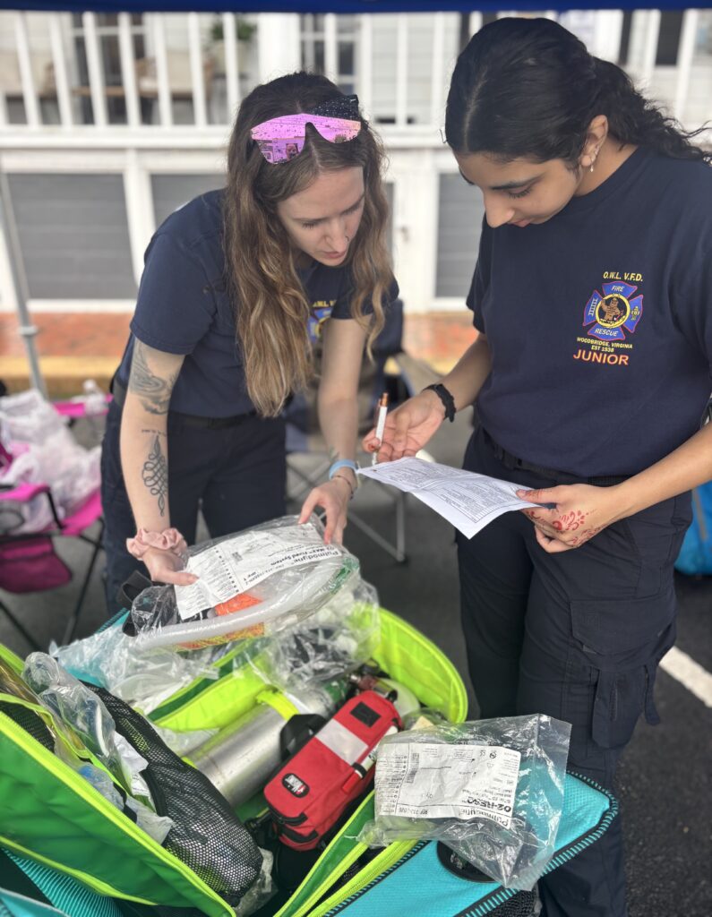 Two young women in uniform review a checklist beside an open medical kit containing equipment and supplies at what appears to be an outdoor training or emergency response event.