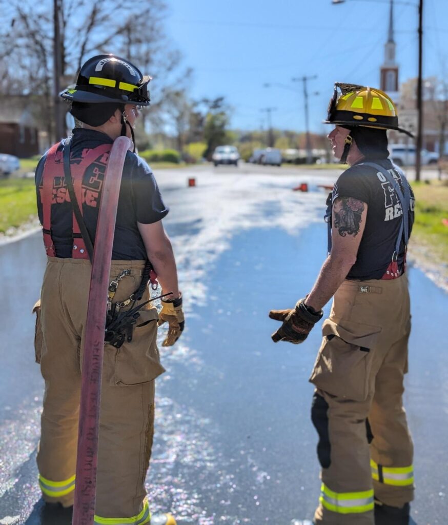 Two firefighters in helmets and gear stand on a wet road, facing away from the camera. One holds a hose over their shoulder as they talk. Sunlight reflects off the pavement, and a church steeple is visible in the background.